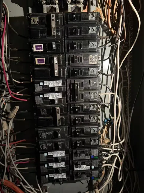 Older residential electrical panel with the cover off, showing a crowded row of breakers and wiring before replacement